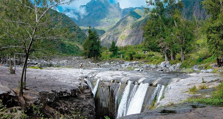 Water stort zich naar beneden over een meerlagige waterval in een groene bergamfitheater, met een enkele wandelaar als schaal voor vergelijking.