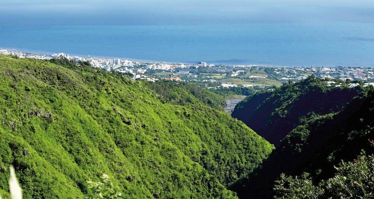 Las paredes del cañón verde y pronunciadas descienden hacia una ciudad costera y el océano azul a lo lejos.