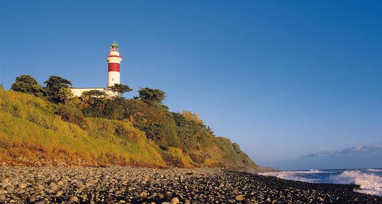 Un faro rojo y blanco se alza en lo alto de un acantilado exuberante sobre una playa de guijarros y olas que rompen.