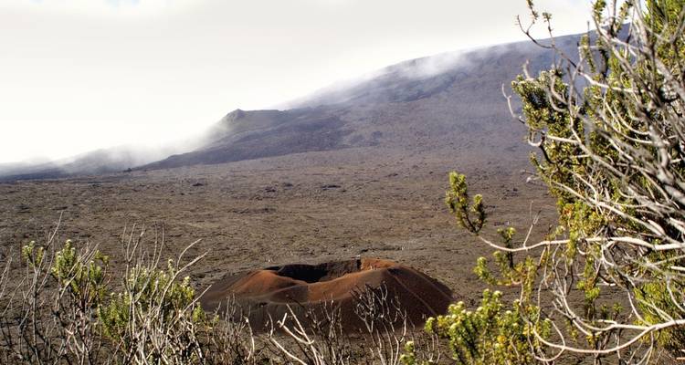 Vista hacia un pequeño cráter volcánico rodeado de campos de lava áridos y colinas brumosas.