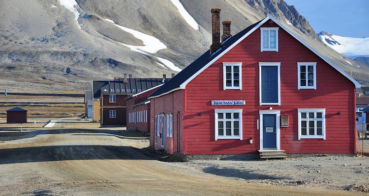 Un edificio tradicional de madera roja se encuentra en una calle de grava con picos árticos escarpados alzándose detrás.