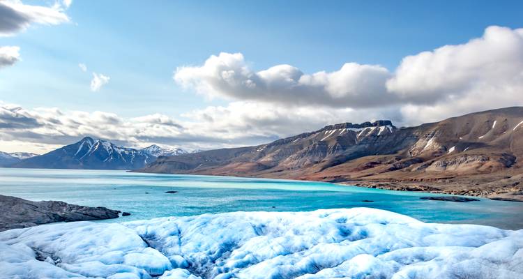 Un glaciar azul vívido conduce a aguas turquesas del Ártico enmarcadas por montañas escarpadas multicolores y nubes dramáticas.