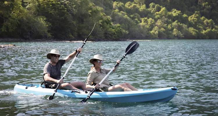 Deux voyageurs pagaient dans un kayak bleu à travers des eaux tropicales calmes avec des collines verdoyantes luxuriantes en arrière-plan.