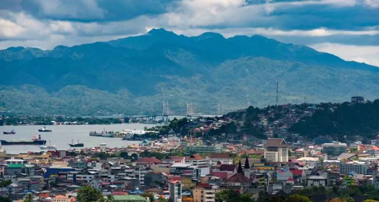 Vue panoramique sur une ville côtière indonésienne avec un pont, un port et des montagnes boisées sous des nuages dramatiques.