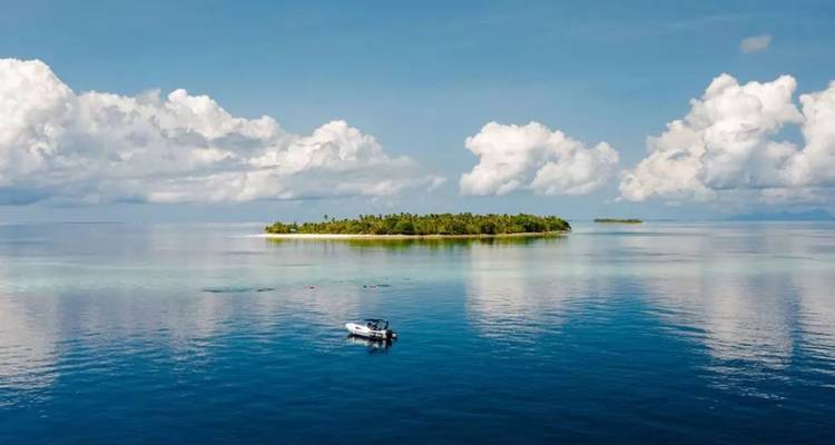 Une minuscule île bordée de palmiers se dresse dans une mer turquoise tranquille avec un petit bateau ancré à proximité sous un ciel bleu.