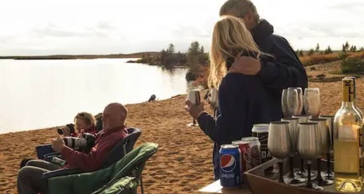 Des voyageurs se détendent avec des boissons et des appareils photo sur une plage de sable au bord d'un lac sous un ciel nuageux.