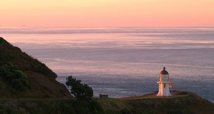 Phare blanc sur un promontoire rocheux dominant l'océan calme sous un ciel de coucher de soleil rose-orange.