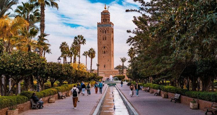 Palmengesäumte Promenade, die zur Koutoubia-Moschee in Marrakesch führt, mit Besuchern, die neben einem langen Wasserbecken spazieren gehen.
