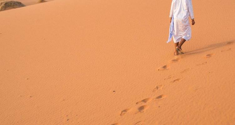 Person in traditionellem Gewand geht allein über orangefarbene Wüstendünen und hinterlässt Fußspuren.