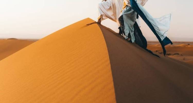 Persoon in wapperende gewaden beklimt steile gouden zanduin onder heldere hemel