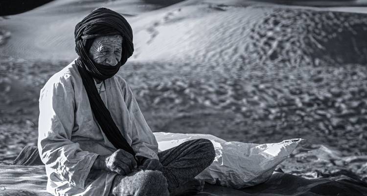Retrato en blanco y negro de anciano habitante del desierto sentado sobre arena con dunas texturizadas detrás