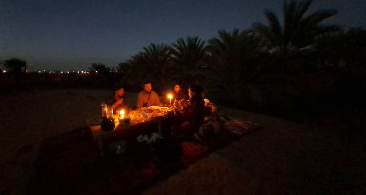 Un pequeño grupo comparte una cena a la luz de las velas sobre una alfombra al aire libre, siluetas de palmeras alrededor, cielo nocturno arriba.