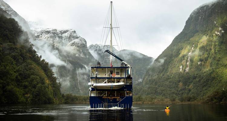 Un navire d'expédition bleu navigue entre des falaises couvertes de forêts tropicales brumeuses et des pics saupoudrés de neige sur un fjord calme.