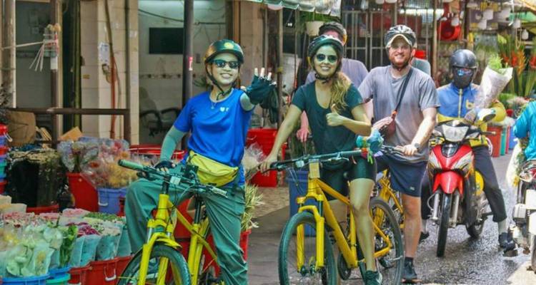 Des cyclistes et un motocycliste traversent une rue animée du marché aux fleurs en agitant la main vers la caméra.