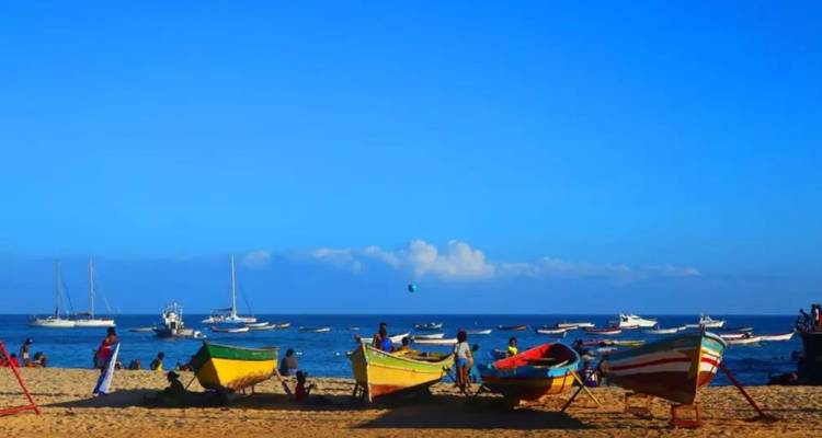 Des bateaux de pêche colorés reposent sur une plage animée tandis que les visiteurs profitent de la mer d'un bleu éclatant sous un ciel dégagé.