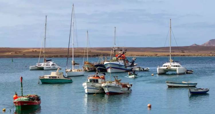De petits bateaux de pêche et de plaisance mouillent dans un port tranquille avec des collines arides au loin.