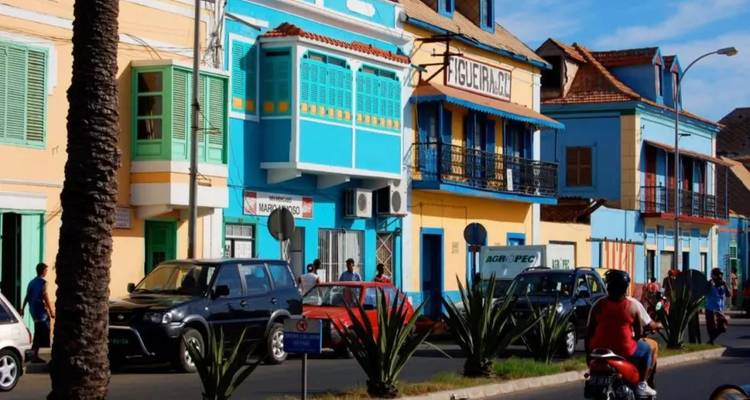 Des bâtiments coloniaux colorés bordent une rue animée du Cap-Vert avec des voitures and des motos qui passent.