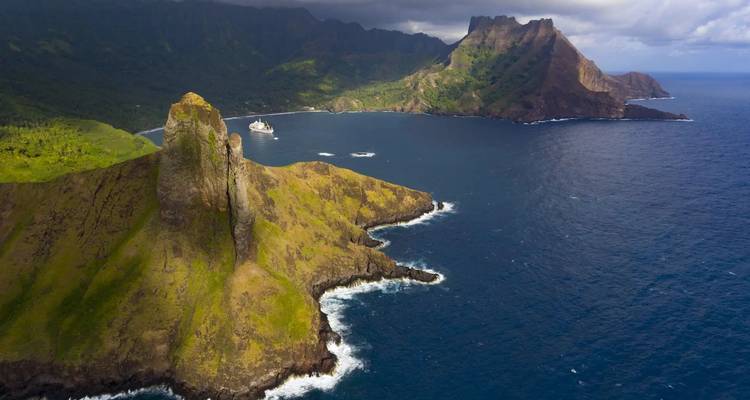 Luftaufnahme der dramatischen Marquesas-Klippen und eines Kreuzfahrtschiffs, das in einer tiefblauen Bucht vor Anker liegt.