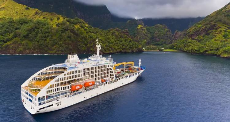 Weißes Passagierschiff vor einer üppigen Vulkaninsel unter düsteren Wolken vor Anker.