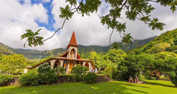 Charmante Kirche mit rotem Dach inmitten eines grünen Tales mit Besuchern davor und nebligen Gipfeln dahinter.
