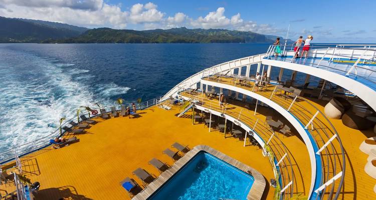 Oberdeck eines Kreuzfahrtschiffs mit Pool und Passagieren, die den Panoramablick auf die Inselberge genießen.