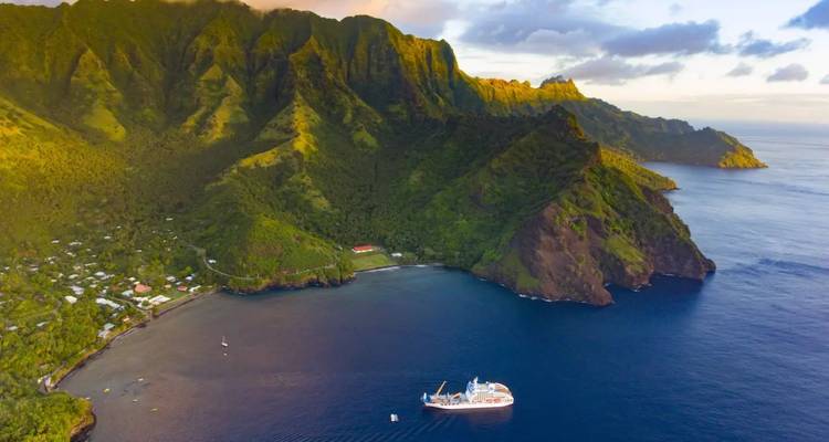 Goldenes Licht taucht steile grüne Bergkämme über einer ruhigen Bucht, wo ein Kreuzfahrtschiff vor Anker liegt.