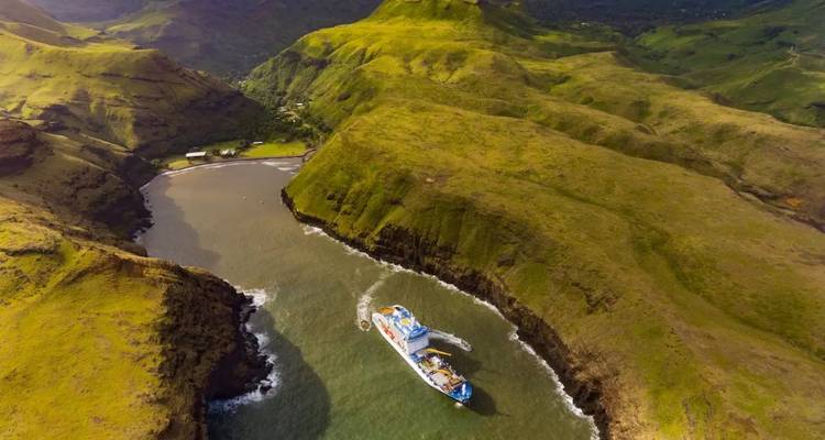 Ein Versorgungsschiff navigiert durch eine schmale, gewundene Bucht, die zwischen grüne Vulkanhügel eingeschnitten ist.