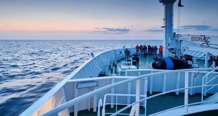 Passengers gather on the bow of a cruise ship at dusk, watching the calm sea and pastel sky.