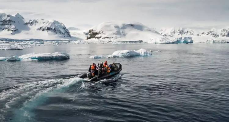 An inflatable zodiac carrying bundled tourists speeds across icy Antarctic waters with snow-covered peaks behind.
