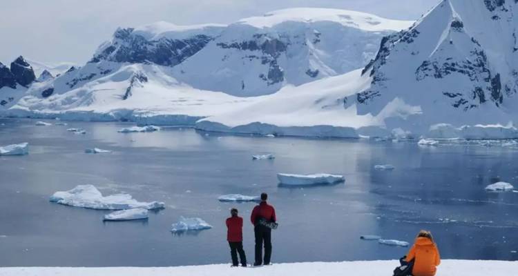 Visitors in colorful jackets stand on snowy ground overlooking an icy bay dotted with ice floes.