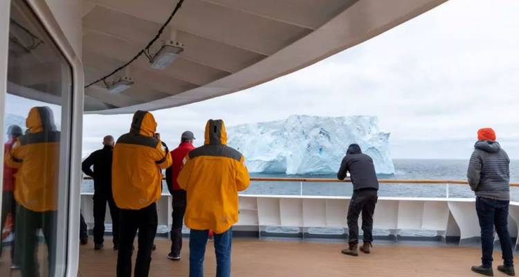 Des passagers en vestes colorées observent un iceberg tabulaire massif depuis le pont d'un navire d'expédition.