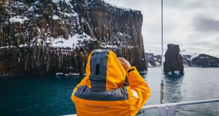 Passager d'expédition en parka jaune photographie des falaises escarpées striées de neige depuis un navire.