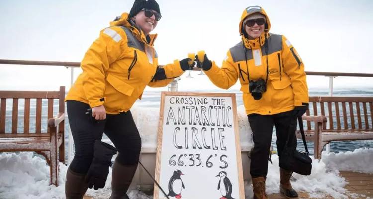 Deux voyageurs trinquent à côté d'un panneau marquant leur traversée du cercle polaire antarctique sur le pont enneigé d'un navire.