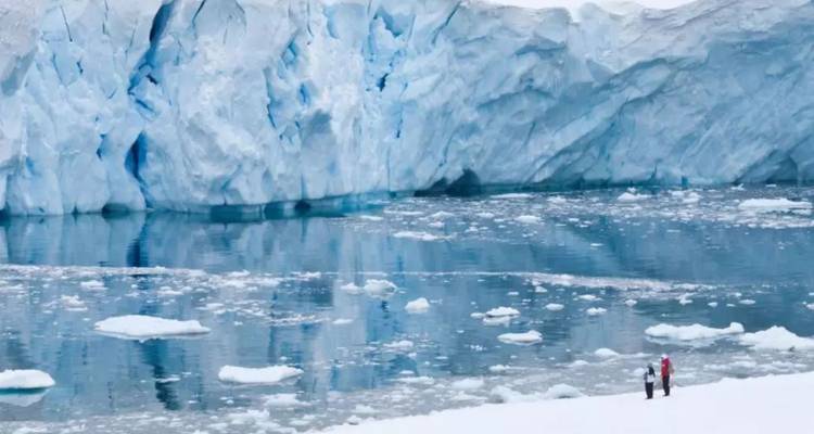 Towering blue-tinged glacier face mirrored in icy water while two small people stand on snowy shore.
