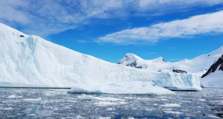 Icebergs and a snowy ridge rise above a calm, floe-strewn sea beneath a bright blue sky.