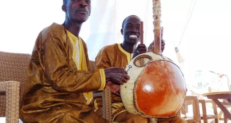 Deux musiciens vêtus de tenues traditionnelles souriant tout en jouant un instrument à cordes de style kora à l'intérieur