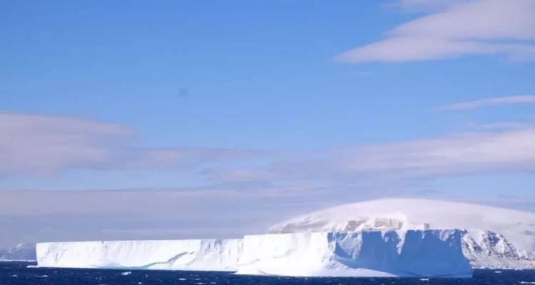 A huge tabular iceberg floats on deep blue water beneath a bright sky.