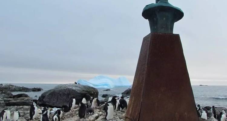 Penguins mill around a rusted beacon on a rocky Antarctic shore with an iceberg beyond.