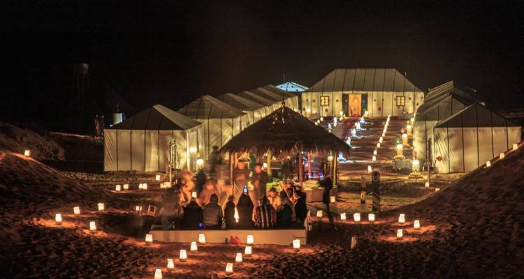 Desert camp lit at night with tents and people gathered around a fire.