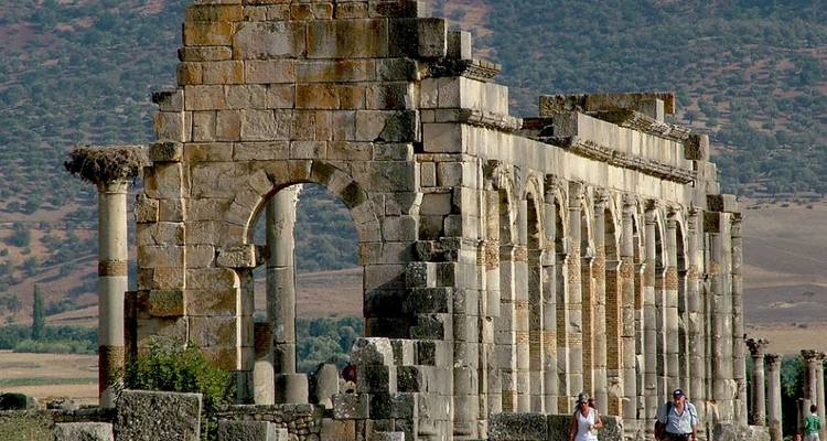 Roman ruins with arches and columns, two people walking near the ruins.