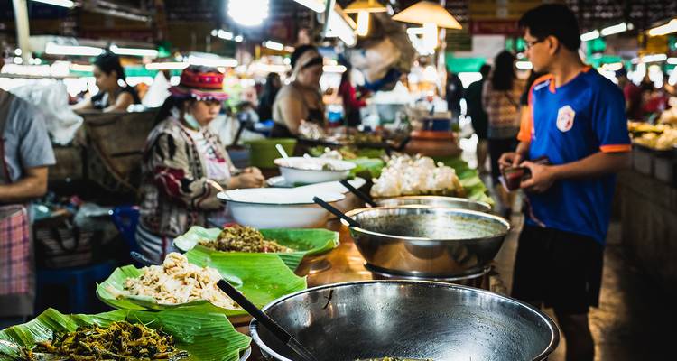 Bustling Thai market food stall with large metal bowls of vibrant dishes and customers ordering.