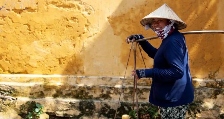 Un vendeur vietnamien souriant portant un chapeau conique transporte des produits contre un mur jaune usé.