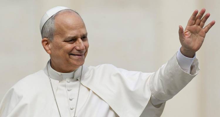 Homme souriant vêtu de robes papales blanches et d'une calotte relevantsa main en signe de salutation.