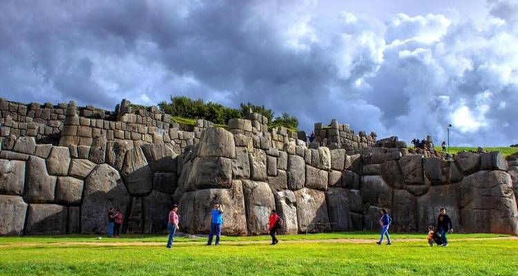 Les imposants murs en pierre de Sacsayhuamán en zigzag inca avec des visiteurs explorant l'esplanade herbeuse.