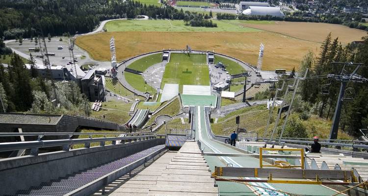 Vista superior hacia abajo de una rampa de salto de esquí hacia un estadio y paisaje rural abajo.