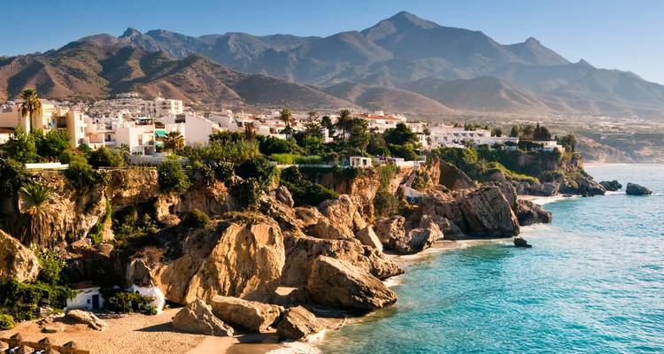 Coastal cliffs dotted with white buildings overlook turquoise Mediterranean waters backed by rugged mountains.
