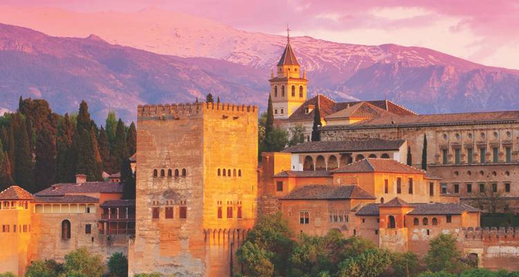 The Alhambra palace complex glows orange beneath snow-capped mountains at sunset.