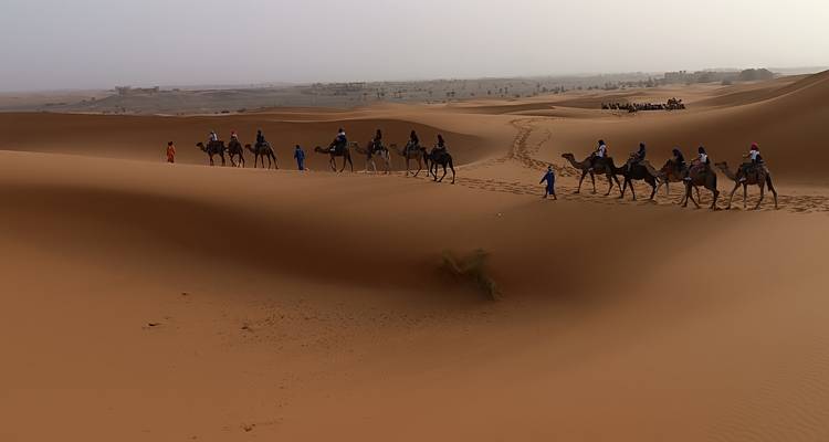 Caravane de chameaux sur les dunes de sable au coucher du soleil avec des gens qui montent.