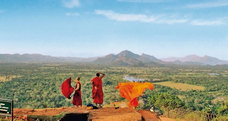 Two monks stand on a rocky outcrop holding saffron robes that billow over a vast valley
