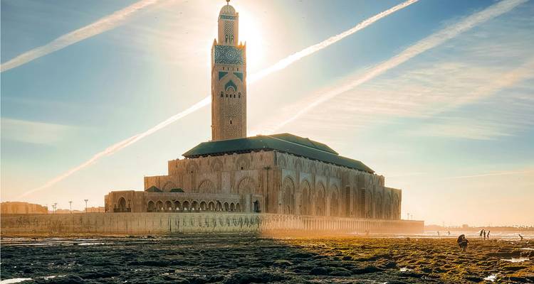 The Hassan II Mosque rises beside the Atlantic Ocean, back-lit by soft golden light with condensation trails streaking the sky.